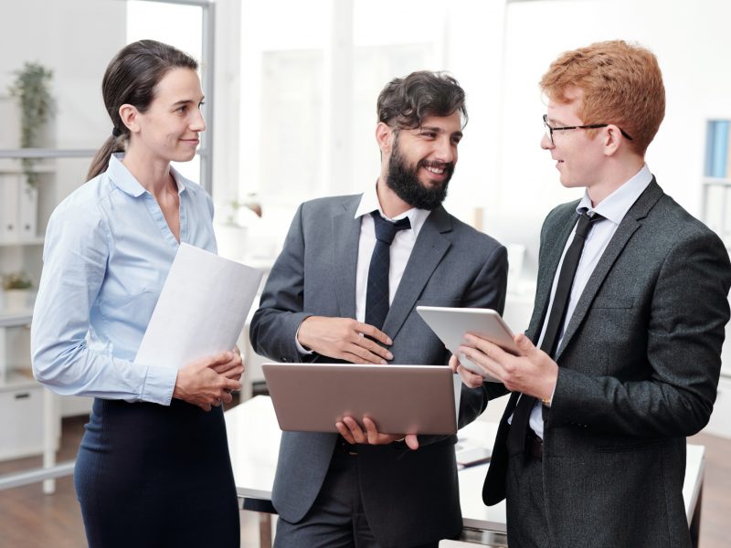 Waist up portrait of three young business people discussing work and smiling while standing in modern office, copy space