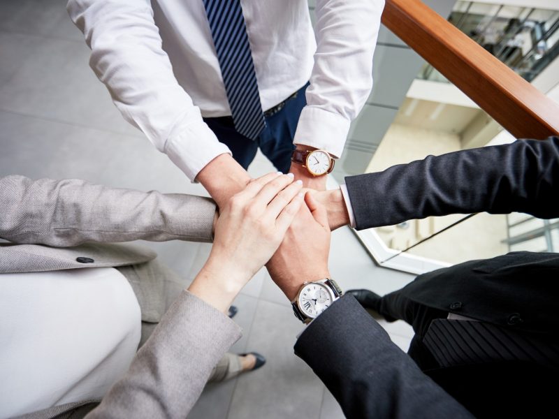 Close-up shot of ambitious team of managers joining hands together after successful completion of negotiations with business partners, interior of office lobby on background