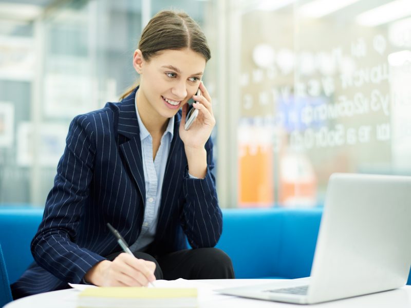 Portrait of smiling young businesswoman speaking by phone in office during internship, copy space